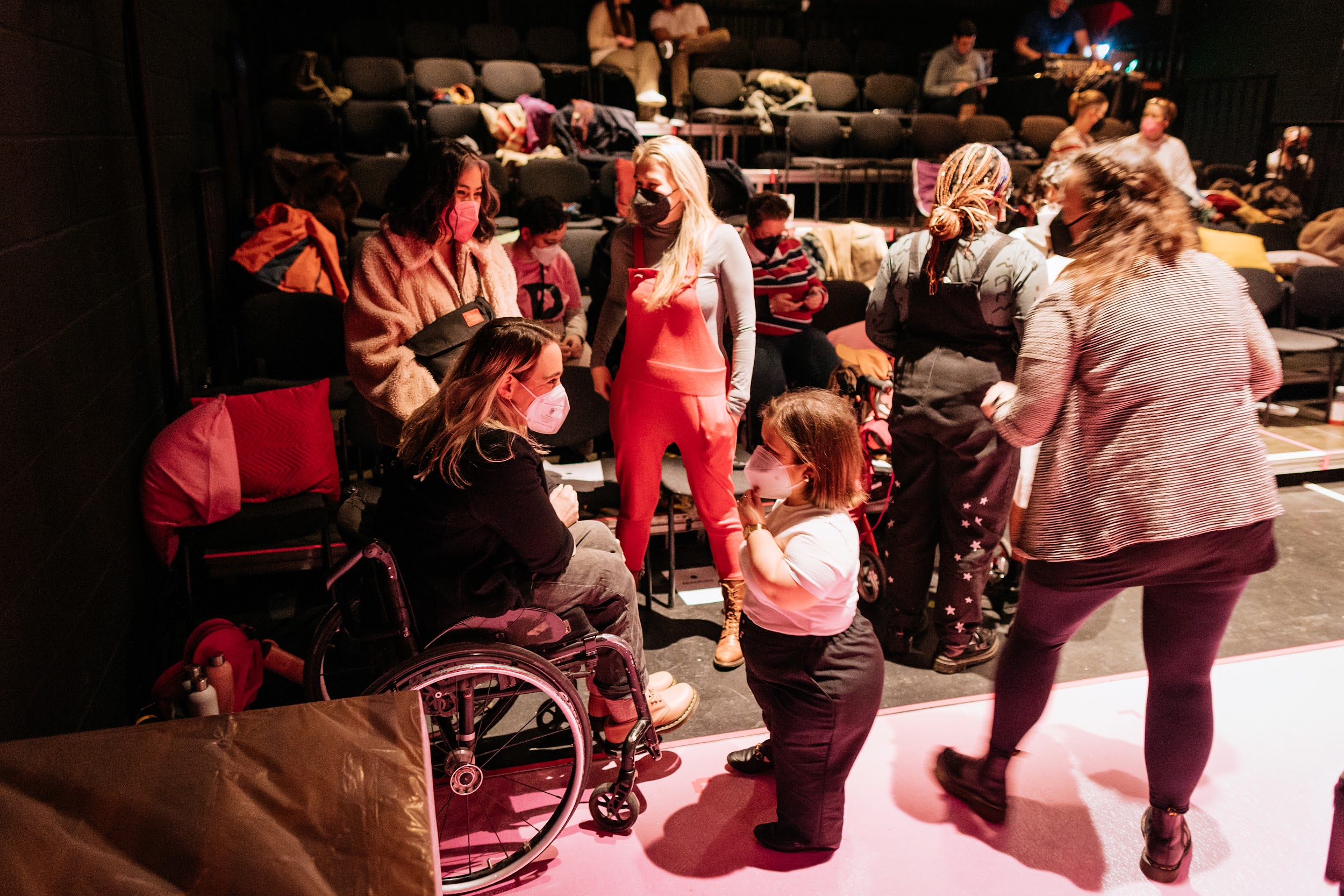 In a candid scene, writer Louise Bruton engages Sinead Burke in conversation. Both people wear masks and are captured in front of the tiered seating of a busy theatre space. Louise Bruton has blonde hair, wears a black jumper and is a wheelchair user. Sinead Burke wears black pants, a white t-shirt and is short in stature. Photo by Simon Lazewski.