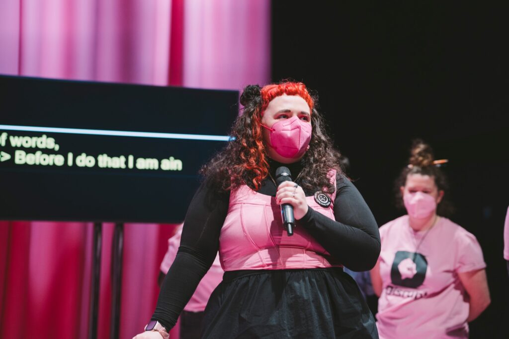 Disrupt festival organiser Áine O'Hara stands on a stage, speaking into a microphone. They wear a facemask. Their hair, clothing and background are all in black and pink. Behind Áine a screen displays real-time text of their words. Photo by Leanne Sullivan.