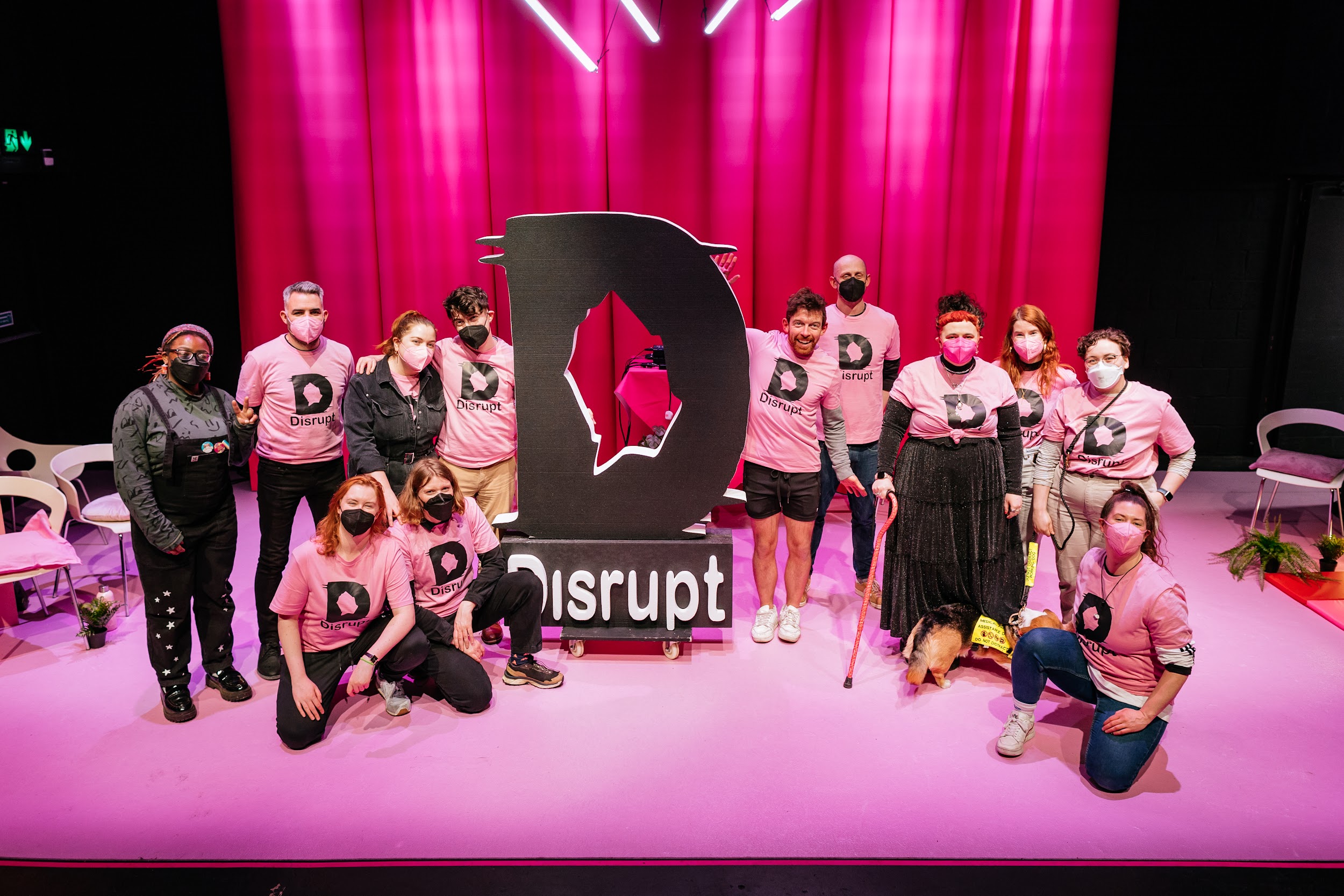 The Disrupt festival stage. Around the Disrupt styrofoam logo the disrupt crew pose. All are dressed in pink festival t-shirts and all joyful in demeanour. The set dressing, Carpet and curtain are in hues of pink. Photo by Simon Lazewski.