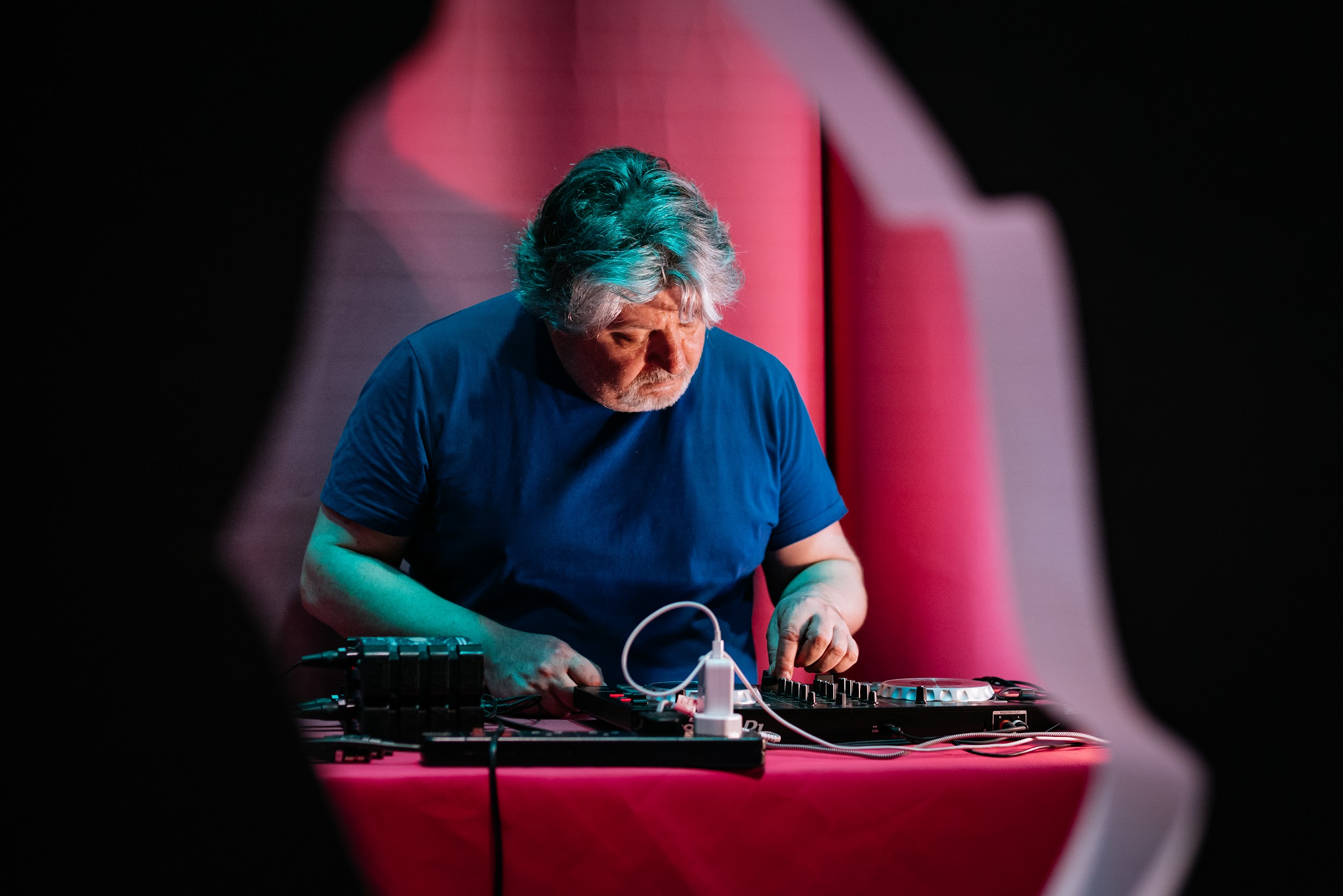 On the Disrupt Disability festival stage is a person in a blue t-shirt with salt and pepper hair. They are sound theatre and sound engineer Jack Cawley They are standing behind DJ equipment, testing it. They are alone; it appears to be pre-show . The image is taken through the central cut out of the styrofoam Disrupt branding. This makes it appear that Jack is framed by a shard of broken glass.