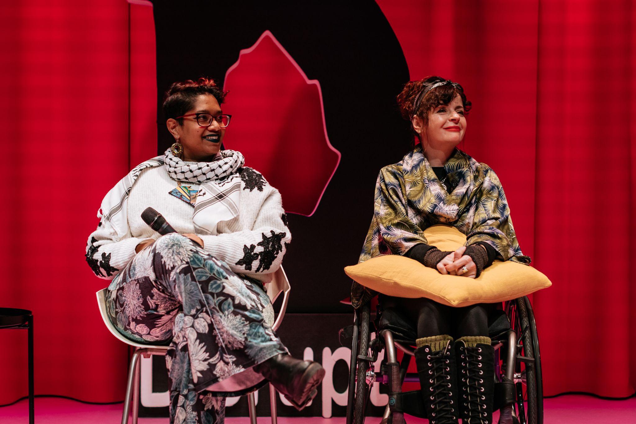 Two people sit facing forward. On the left, a person with short dark hair and dark skin holds a microphone on their lap while seated on a chair. Beside them, a person with dark hair and light skin sits in a wheelchair, with hands resting on a yellow pillow on their lap. Both have heads turned slightly to the left and are smiling. Behind them is a vibrant pink background with a large black letter “D” for Disrupt Disability Arts Festival. Photo by Simon Lazewski.