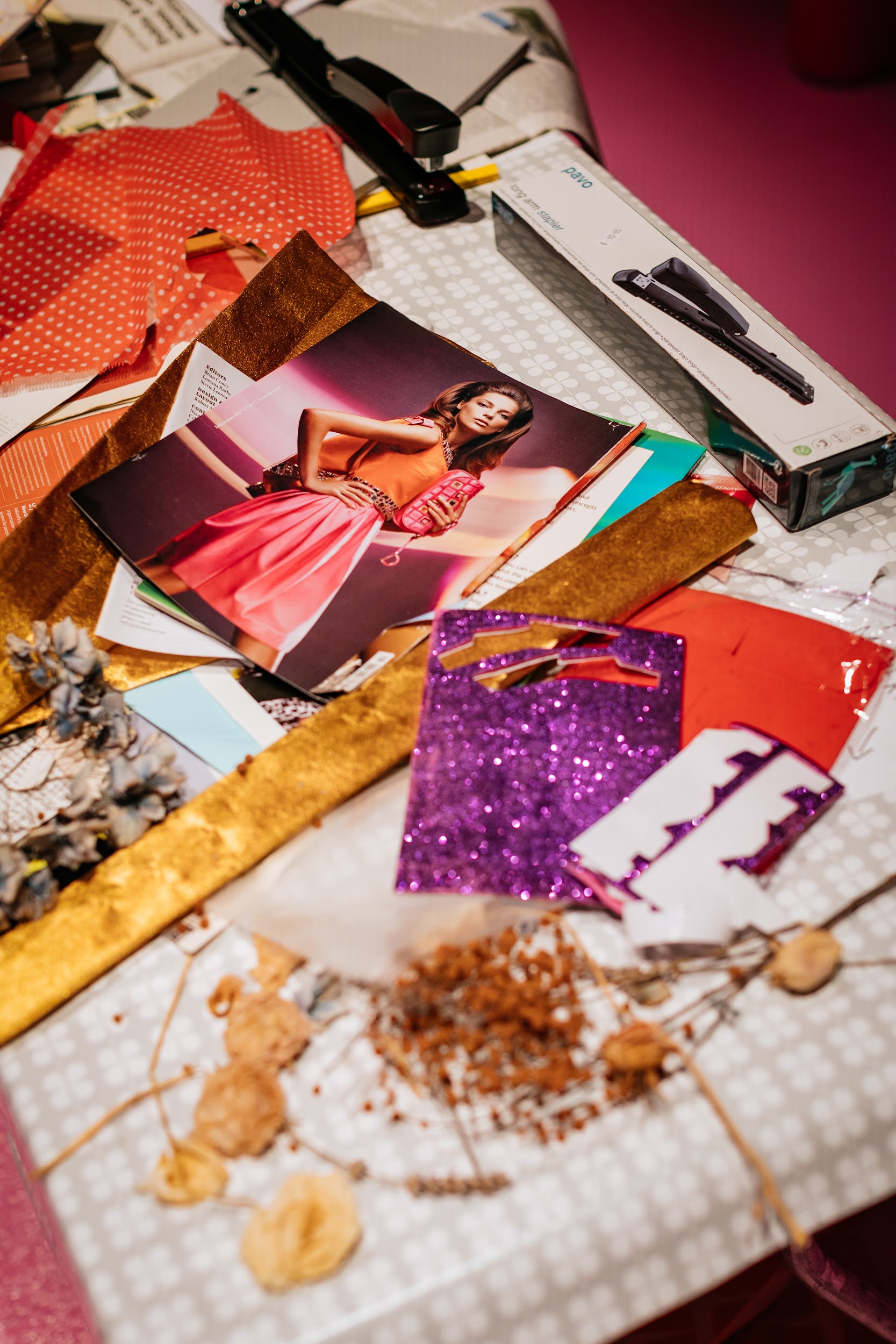 An ad-hoc collection of material upon a tabletop. Items include a stabler, coloured and glitter paper. Fashion magazines and dried flowers. Photo by Simon Lazewski.