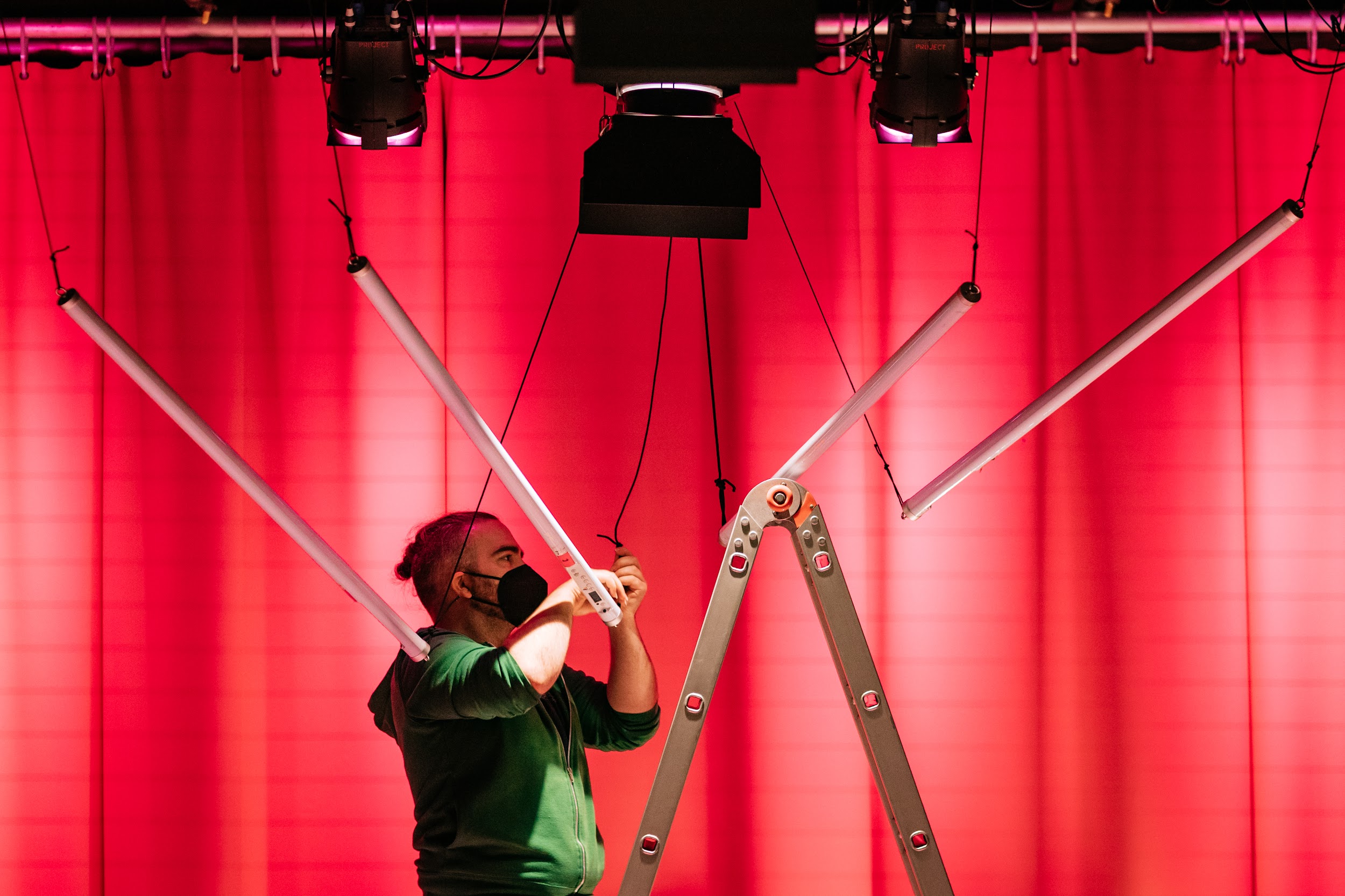 Lighting designer Eoin Lennon is up a ladder in front of the Disrupt Disability Arts Festival stage curtain. Eoin wears a green hoodie and a black face mask. He is partially spotlit. Eoin is wiring a tube light which hangs above the stage from the rafters. The light is one of four which all hang at angles to point down towards centre stage.