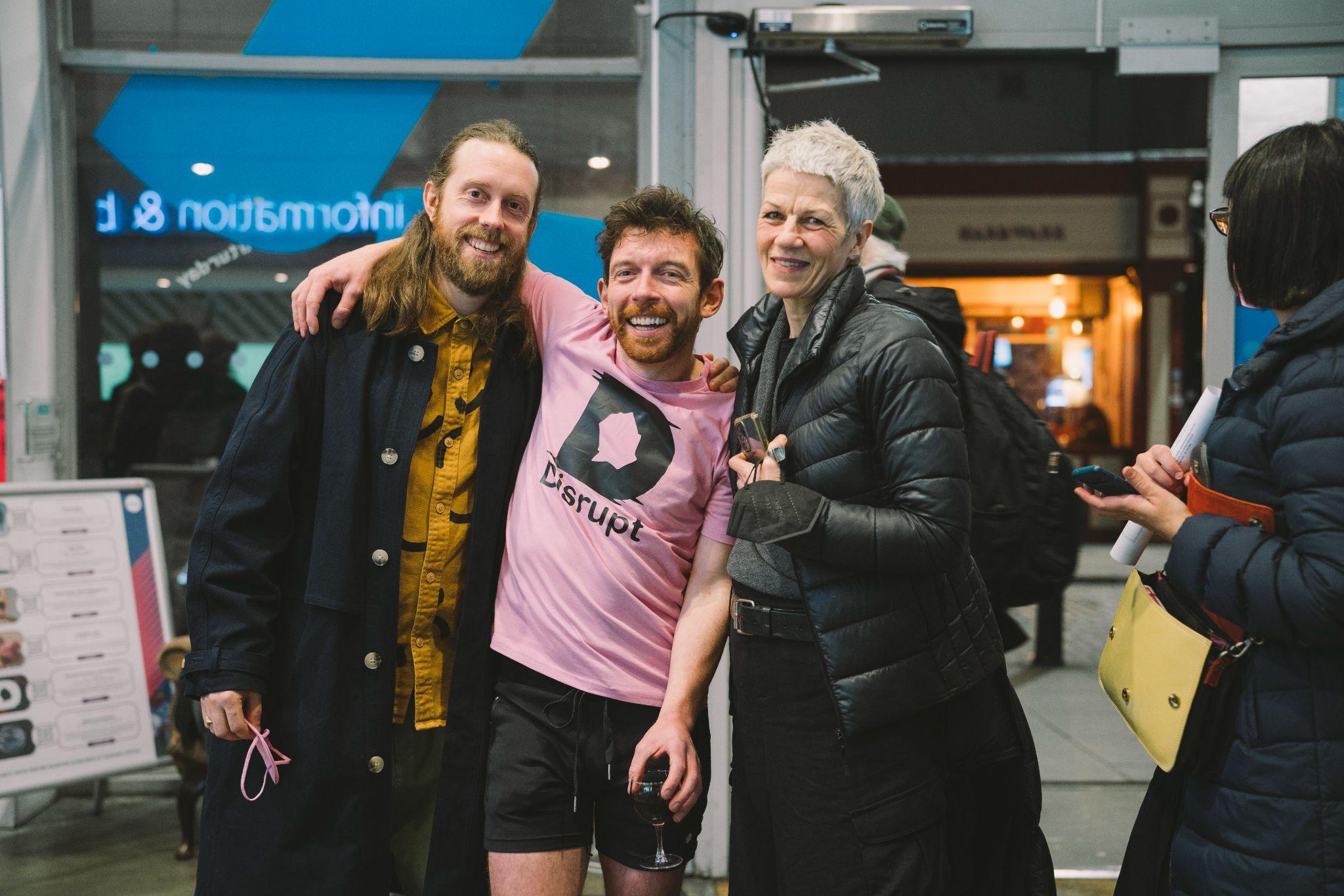 The image shows artist and curator AlanJames Burns standing with two people on either side, all facing the camera and smiling with arms around each other. Alan is dressed in a pink t-shirt featuring the Disrupt Disability Arts Festival logo. All individuals have pale skin. To the right, a person walks into the frame. In the background and out of focus, an open door reveals a busy city street at night. Photo by Leanne Sullivan.