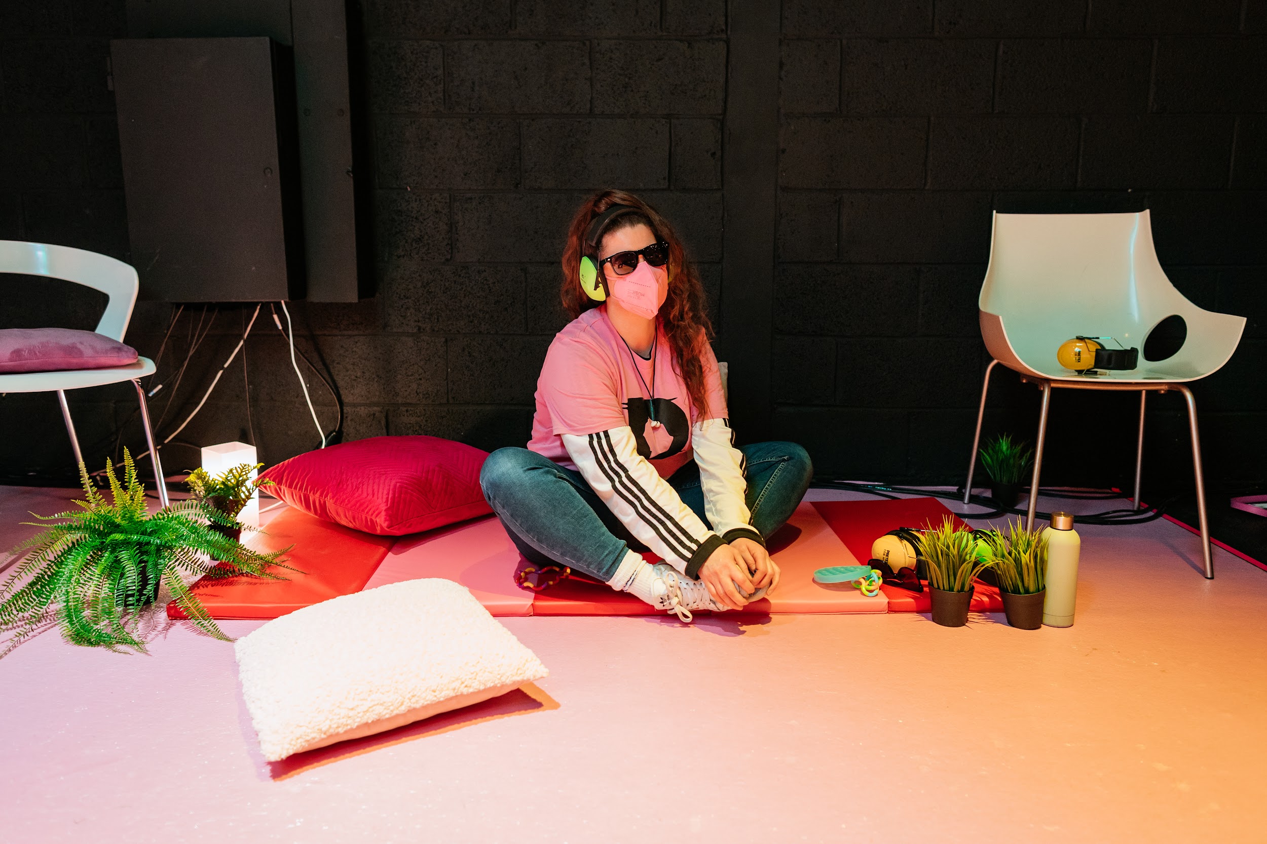 The image shows a person with feminine features sitting on a mat in an indoor location. They wear sunglasses and ear defenders. Around them are plants, cushions, atmospheric lighting and stim toys. Photo by Simon Lazewski.